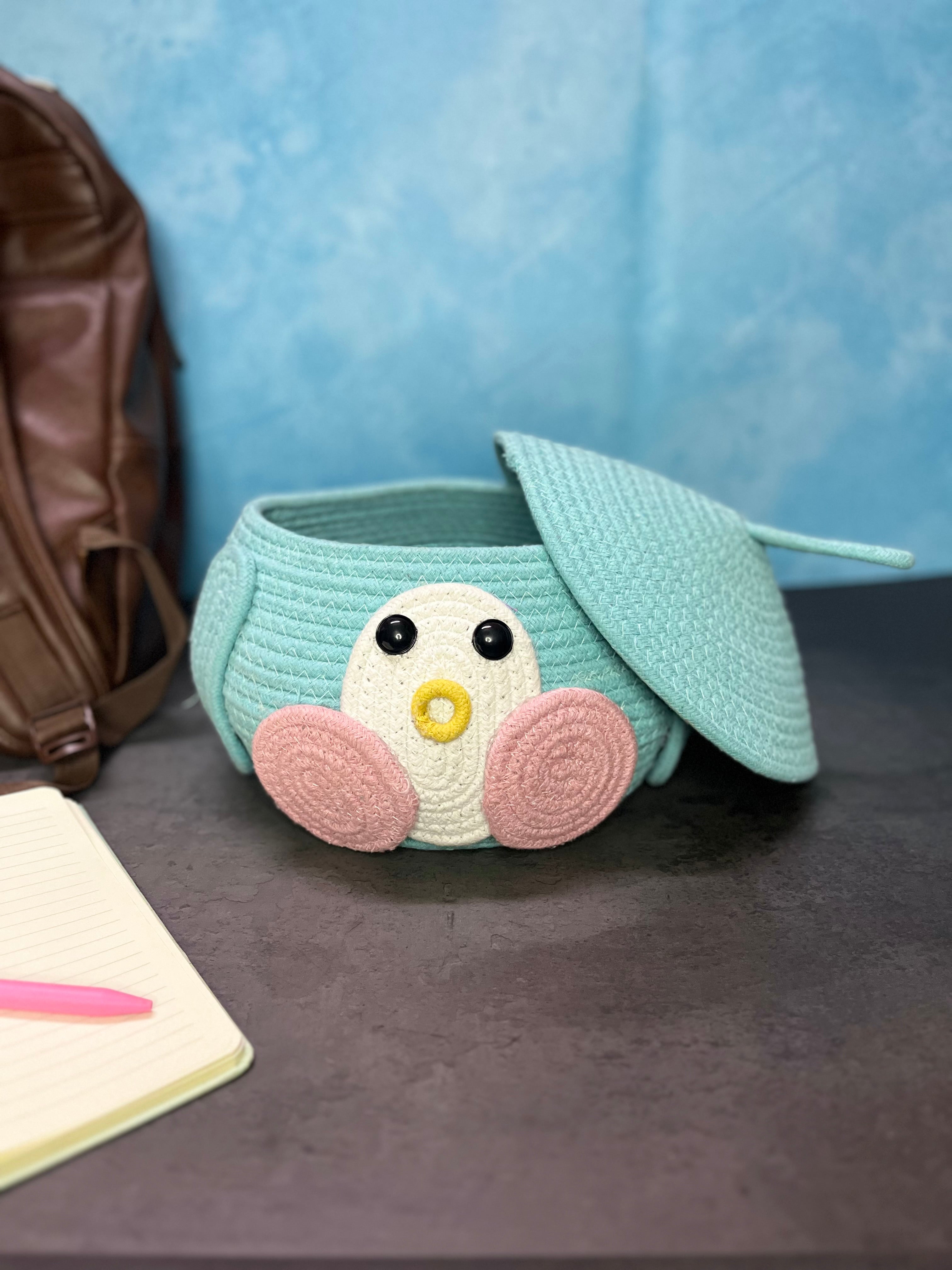 A small, soft woven storage basket in light teal, designed like a cute bird with big black eyes, a yellow beak, and pink cheeks.